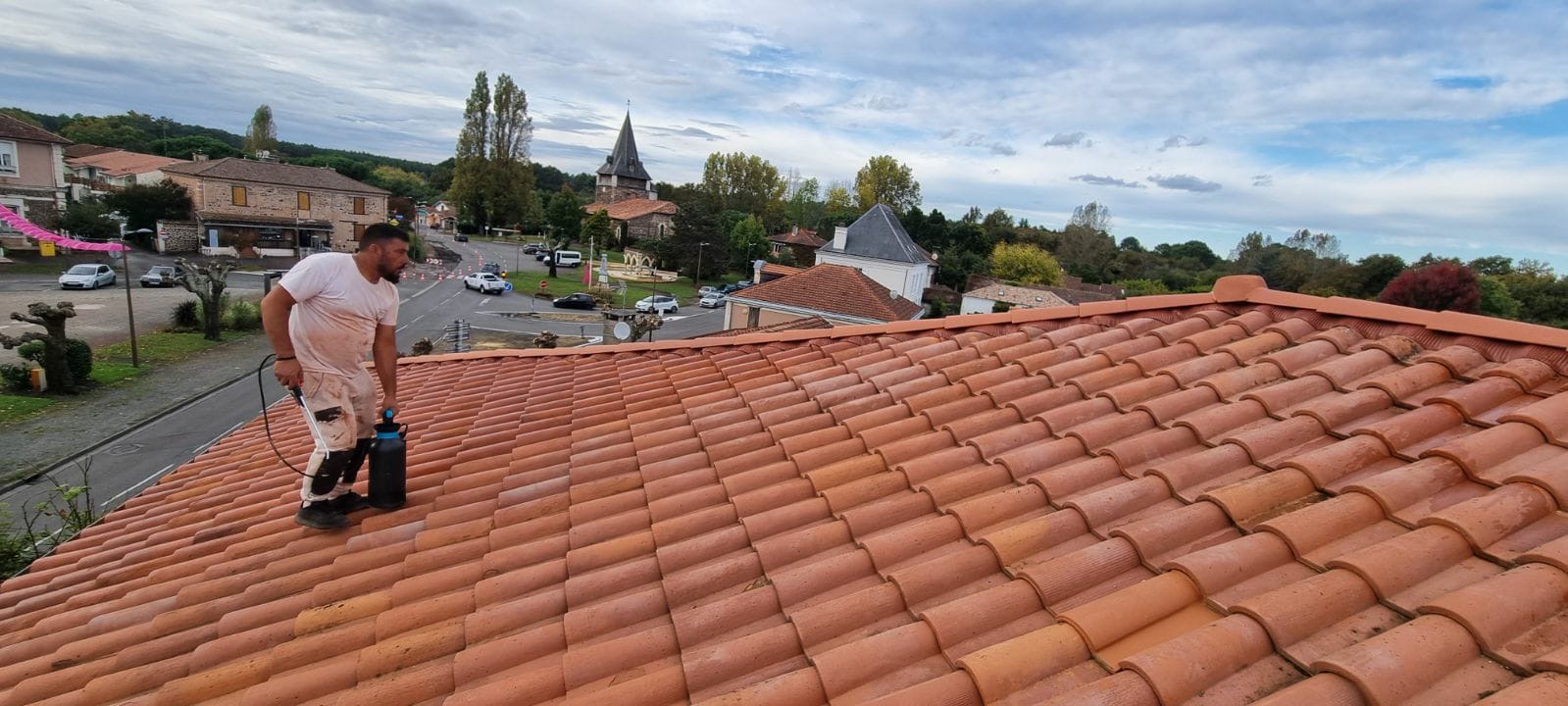 Couvreur appliquant un traitement sur une toiture en tuiles avec vue sur l'église du village en Gironde